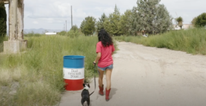 A lady walking with her dog passes right next to a "Don't mess with Texas" trash barrel